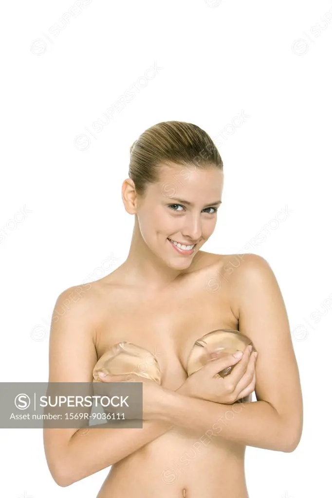 Nude woman holding breast implants over chest, smiling at camera