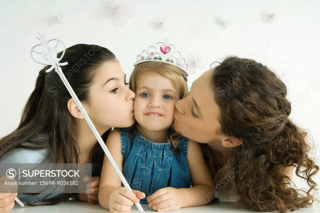 Girl dressed as princess, holding wand, mother and sister kissing her cheeks