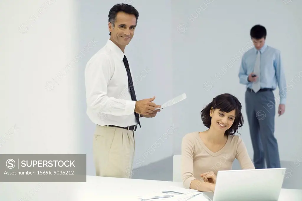 Professionals in office, man standing and holding document, woman sitting in front of laptop computer