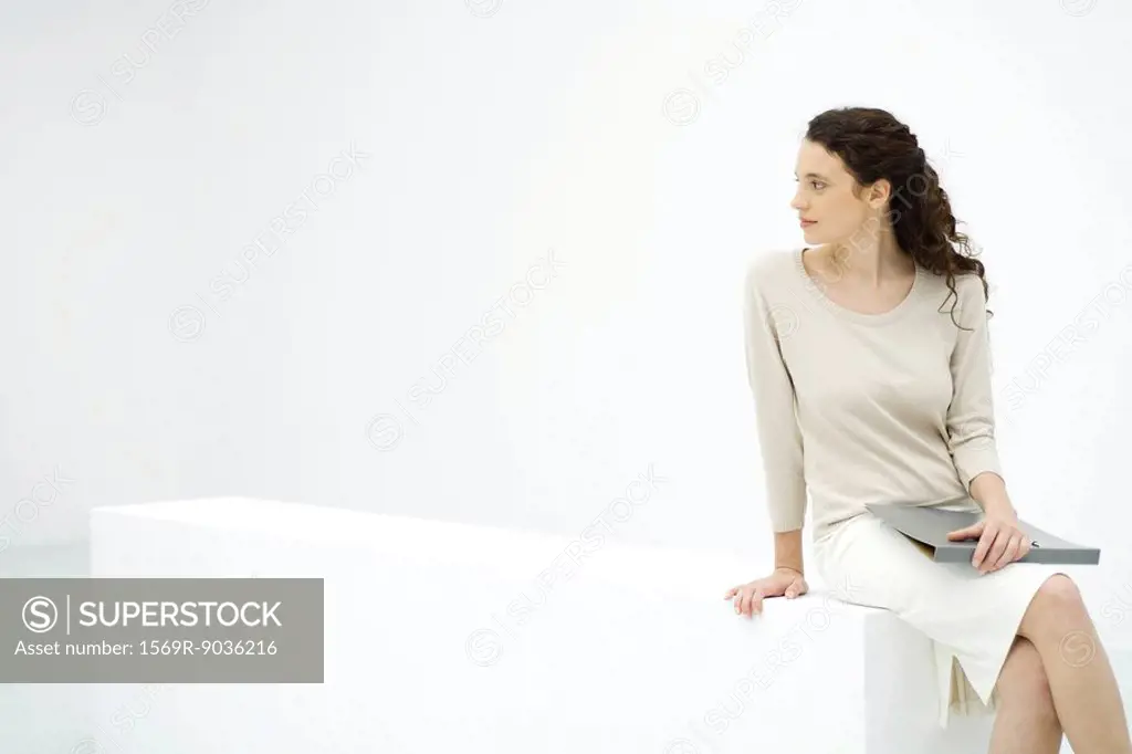 Young female professional sitting on ledge, holding binder, looking over shoulder