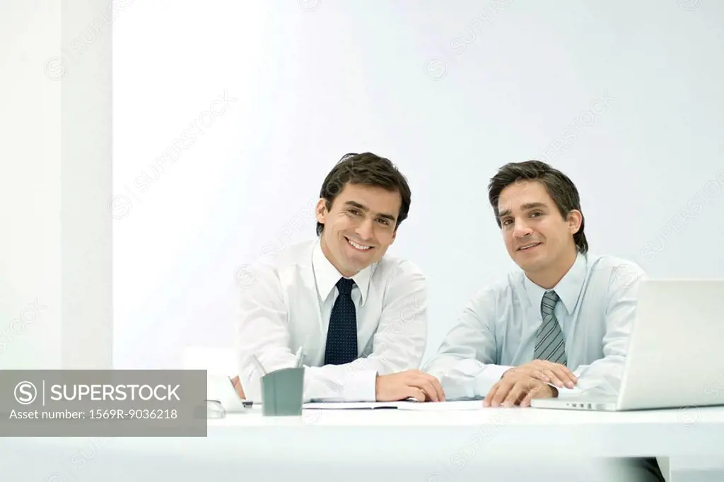 Male business partners sitting side by side at desk, smiling at camera