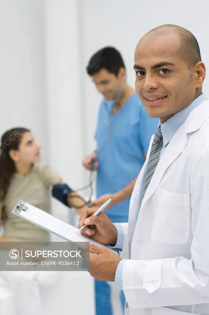 Doctor writing on clipboard, smiling at camera, nurse measuring patient´s blood pressure in background