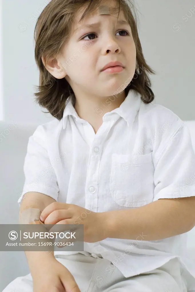 Little boy picking at adhesive bandage on his arm, frowning
