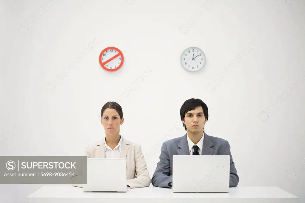Office workers sitting below clocks, using laptops, warning sign over one clock