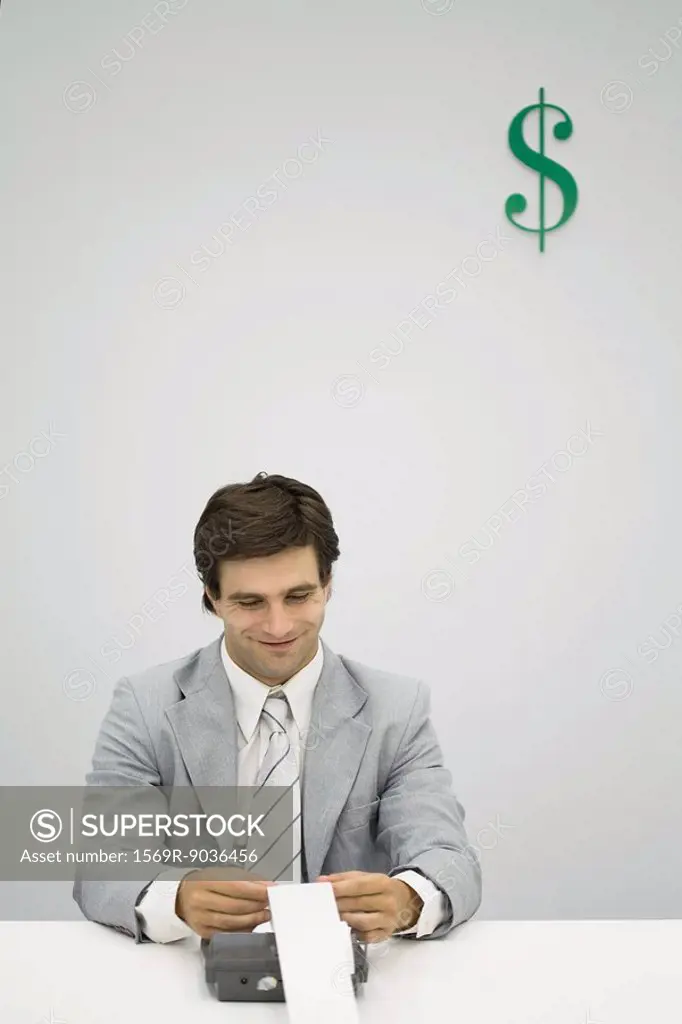 Accountant using adding machine, smiling, dollar sign on wall behind him