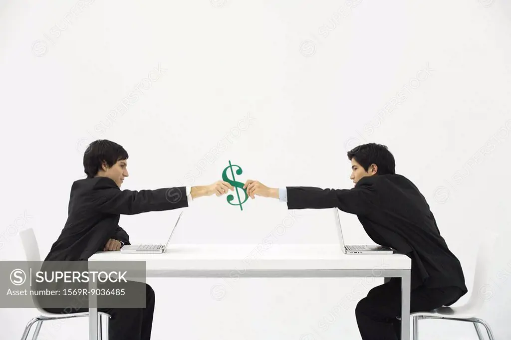 Businessmen sitting face to face at table, each reaching to hold dollar sign