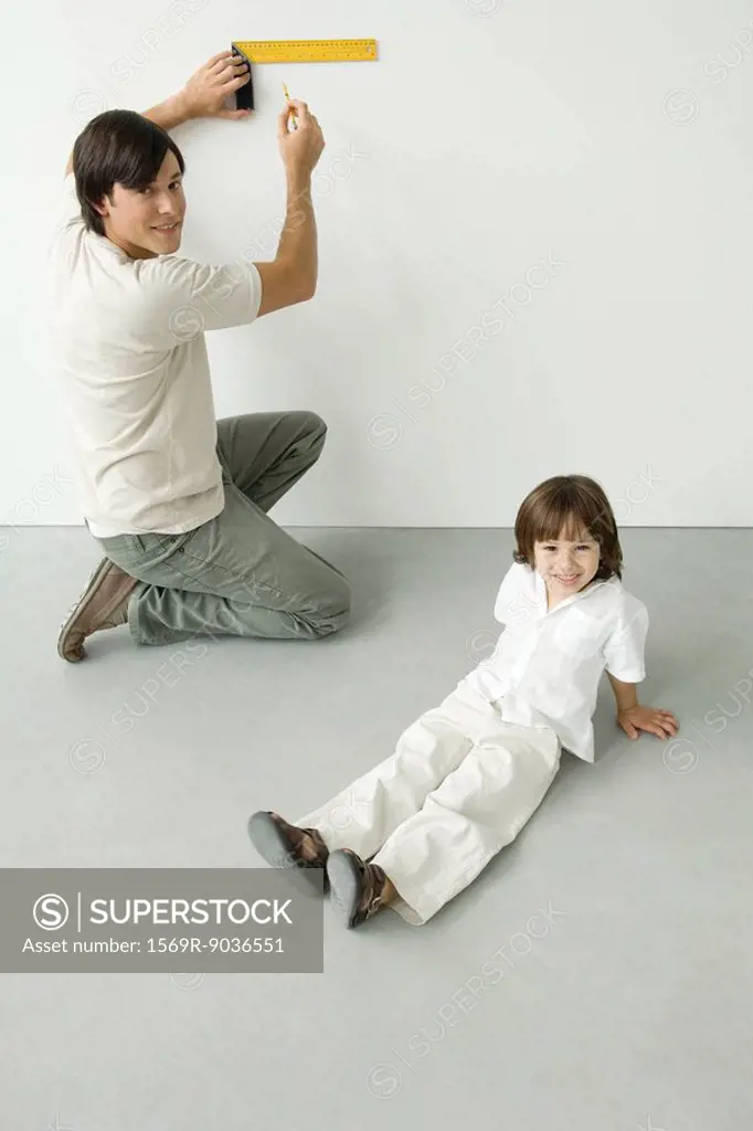 Man measuring wall with a ruler, son sitting nearby, both smiling at camera