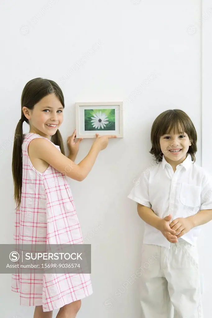 Girl standing beside younger brother, hanging framed picture, both smiling at camera