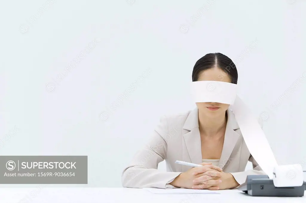 Woman sitting at table with adding machine tape wrapped around her eyes