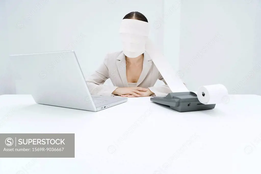 Woman sitting at desk with adding machine and laptop, printout tape wrapped around head