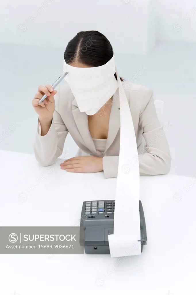 Woman sitting with adding machine tape wrapped around head, holding pen to forehead