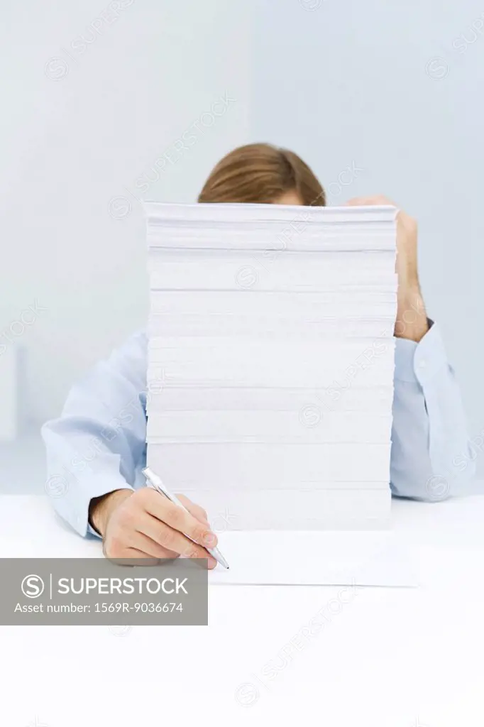 Man hiding behind tall stack of paper, reaching around to write on a single sheet of paper