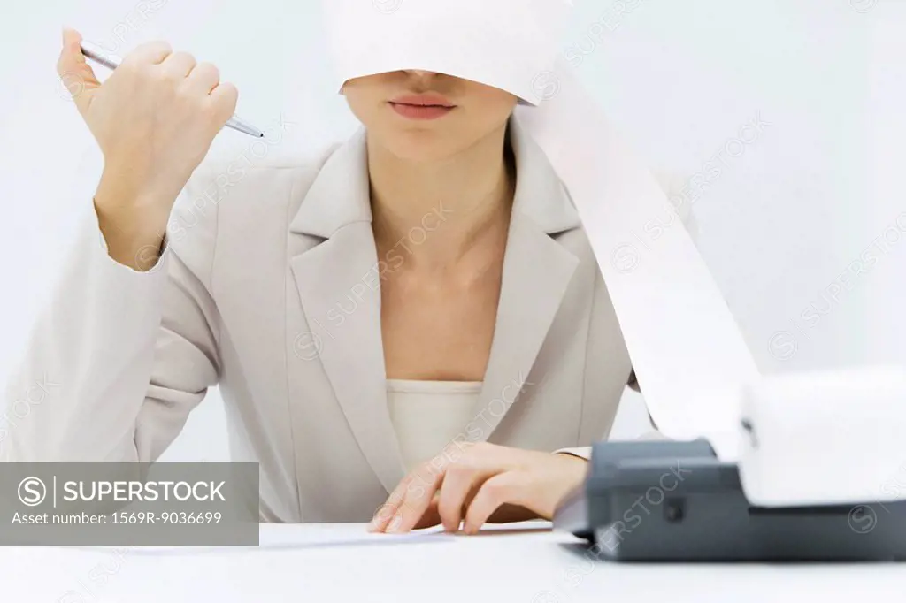 Woman sitting at desk with adding machine tape wrapped around her head, clicking pen