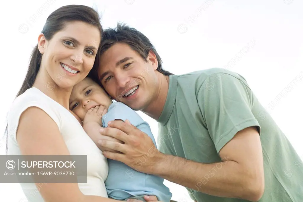 Family smiling at camera, mother holding baby daughter, father leaning in and gently hugging the baby