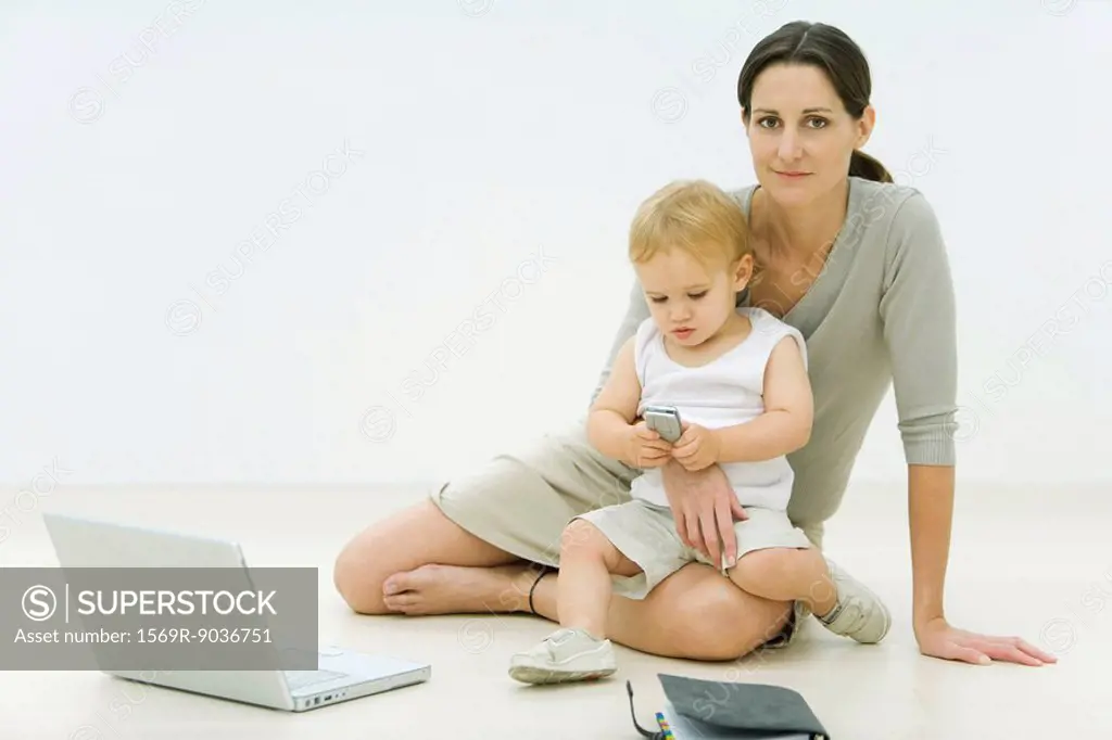 Professional woman sitting on the ground with laptop, holding toddler on lap, toddler looking at cell phone