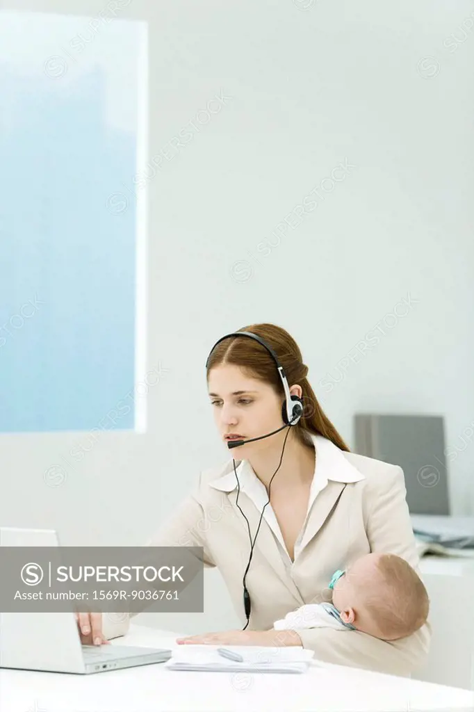 Professional woman in office, holding baby, using headset and laptop computer