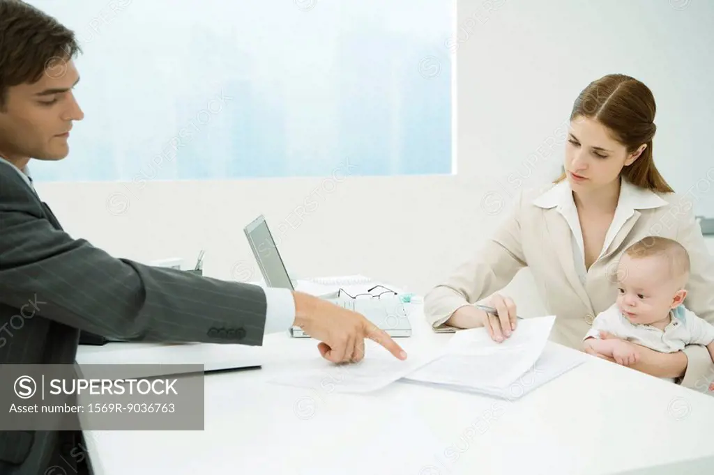 Professional woman in office, holding baby on lap, discussing documents with male colleague
