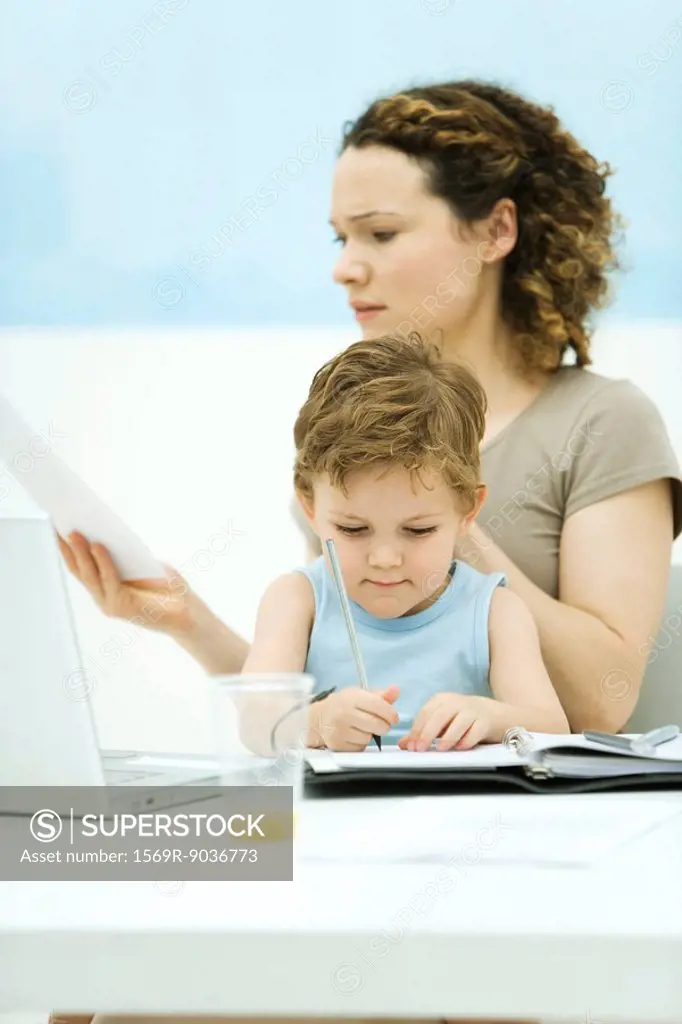 Mother holding son on lap, sitting in front of laptop, frowning at document