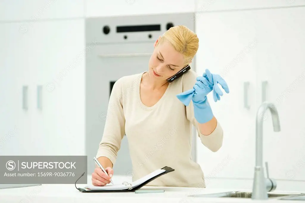 Woman standing at kitchen sink, wearing rubber gloves, using cell phone and writing in agenda