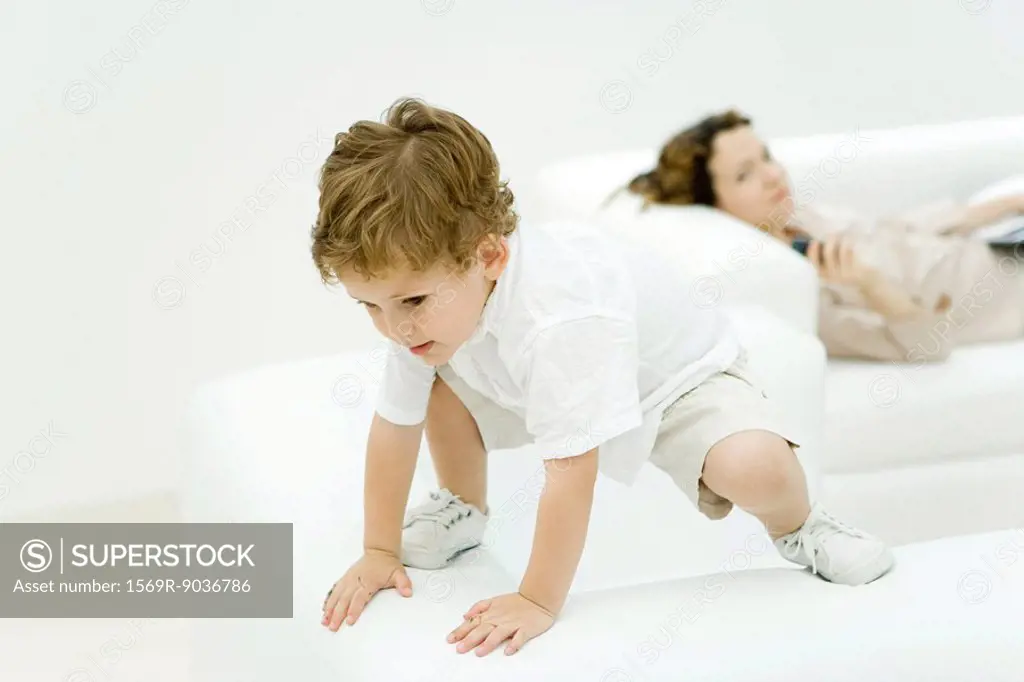 Little boy perched precariously on top of armchair, mother lying on sofa in background