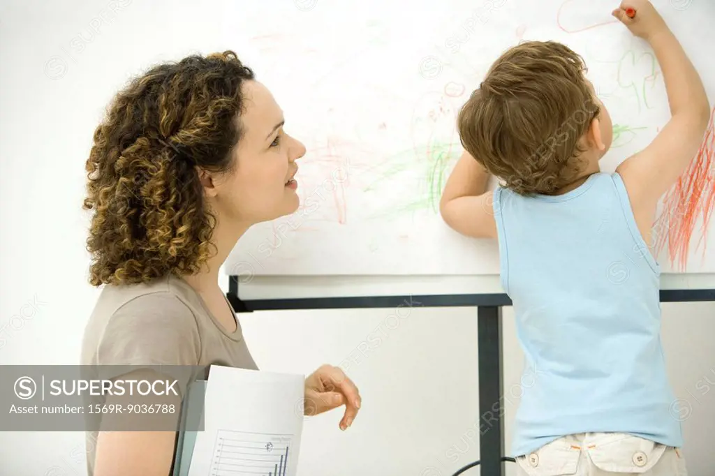 Professional mother holding documents, watching son color with crayon