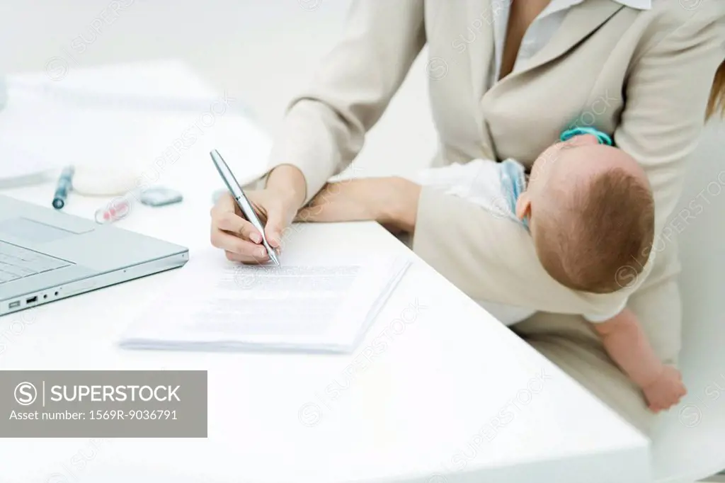 Professional woman signing document at desk, holding sleeping infant, cropped view