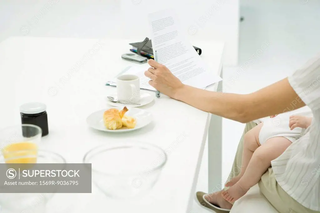 Mother sitting at breakfast table, reading document, infant sleeping on lap, cropped view