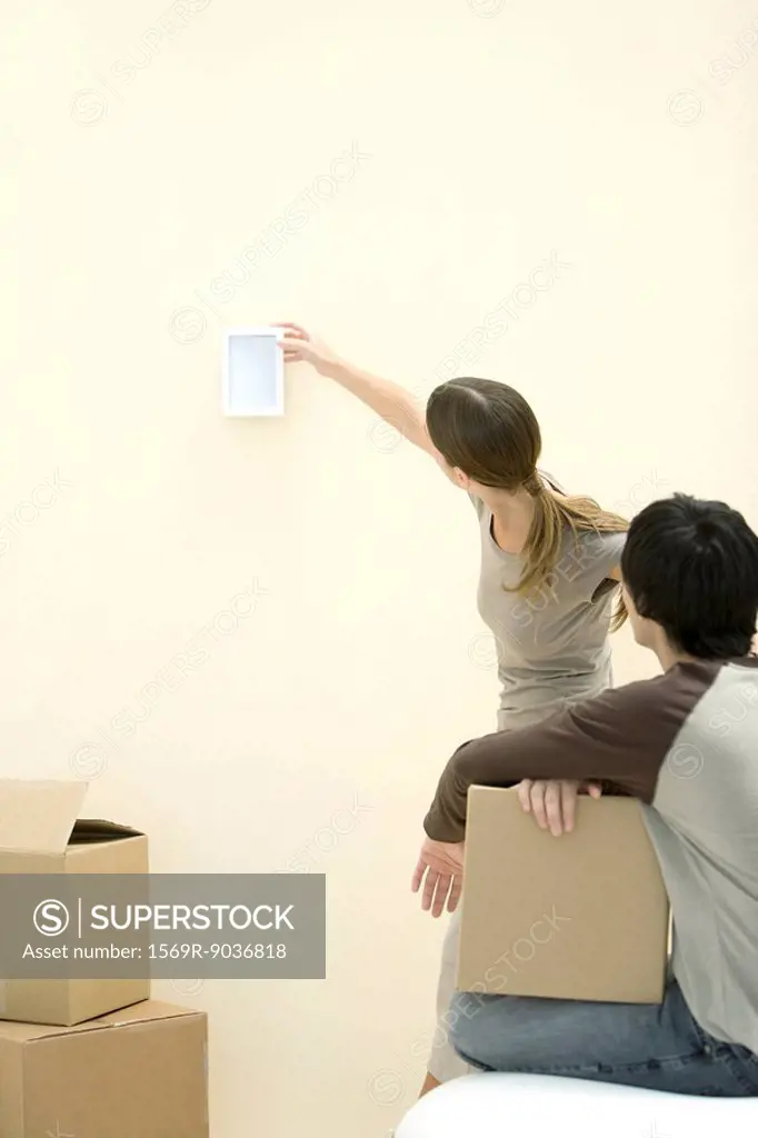 Woman hanging picture frame on wall, man watching, holding cardboard box