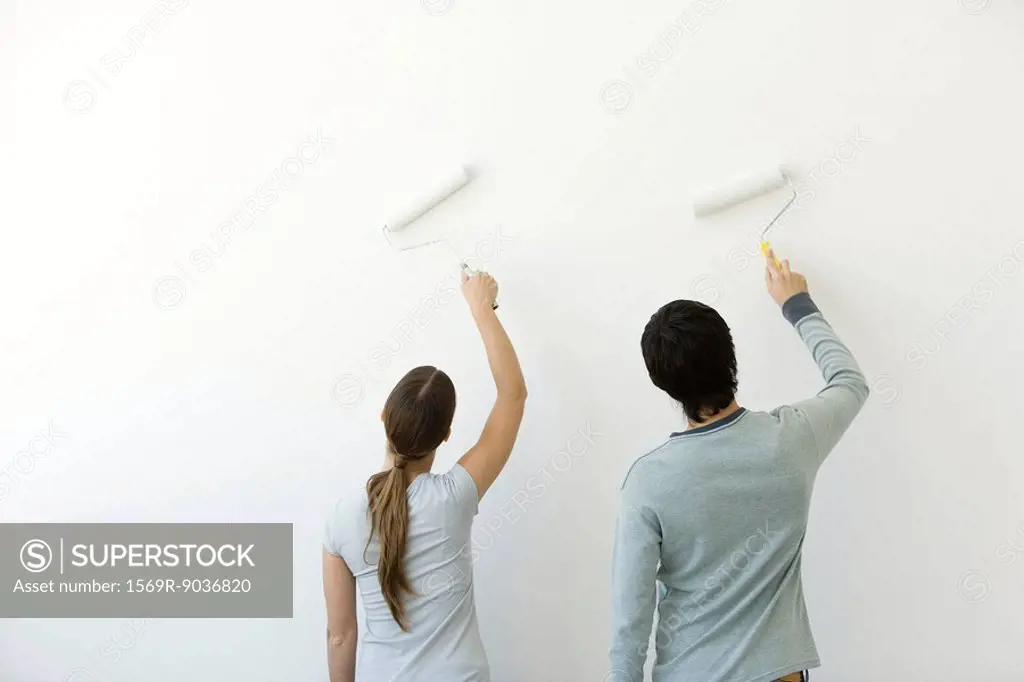 Couple using paint rollers on wall, rear view