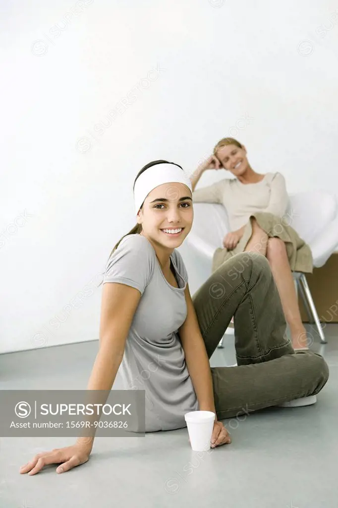 Teen girl sitting on the ground with drink, smiling at camera, mother sitting in background