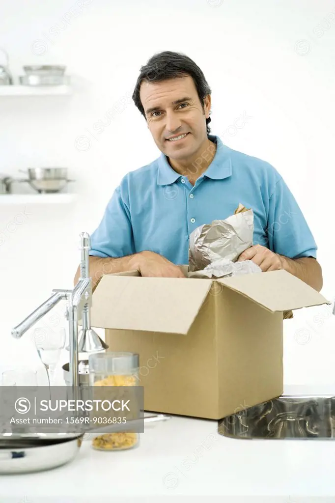 Man unpacking box on kitchen counter, smiling at camera