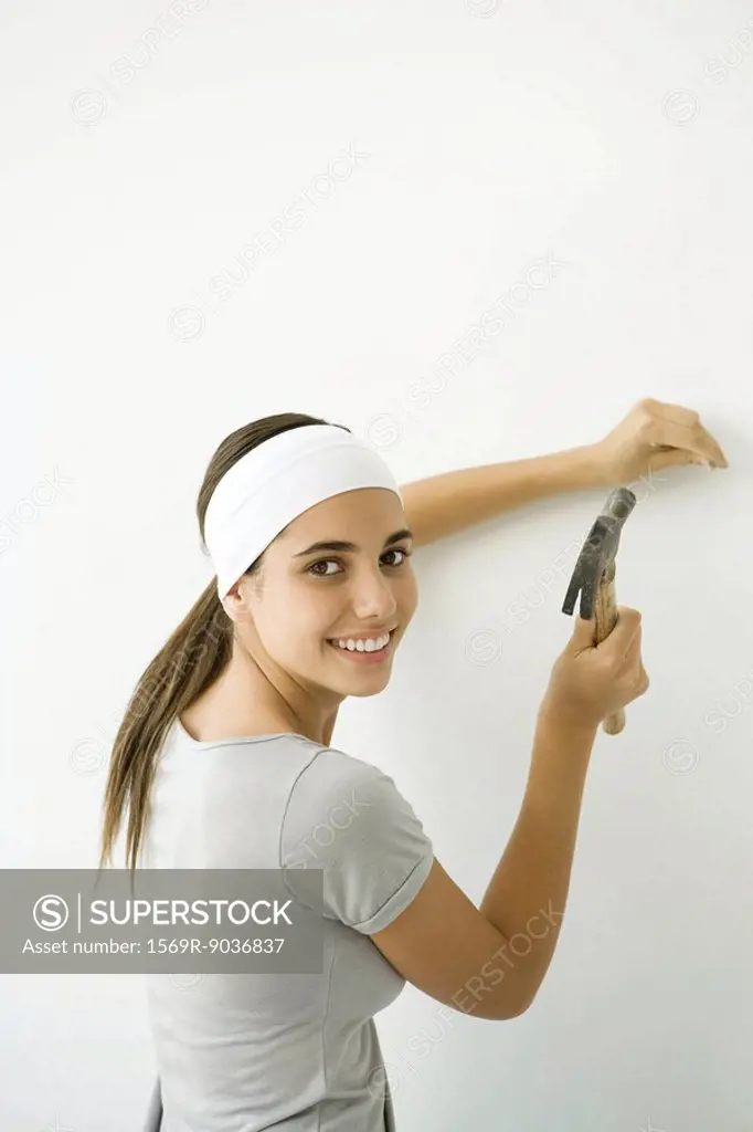 Teen girl hammering nail into wall, smiling over her shoulder at camera