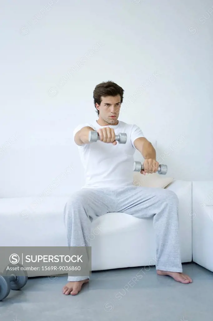 Young man sitting on sofa, lifting dumbbells