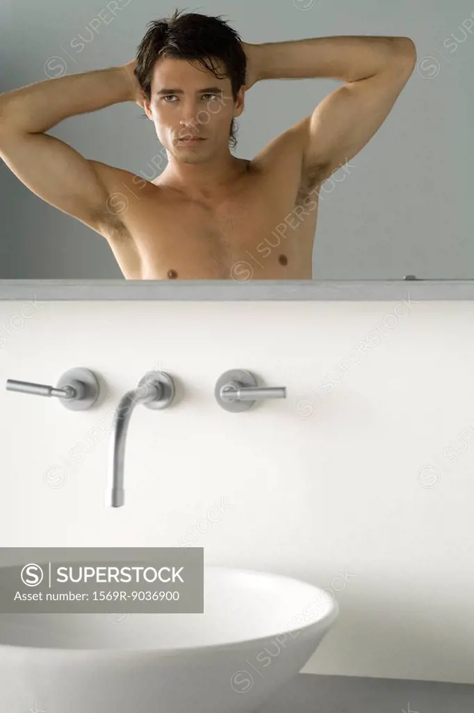 Bare-chested man looking at self in bathroom mirror, hands behind head