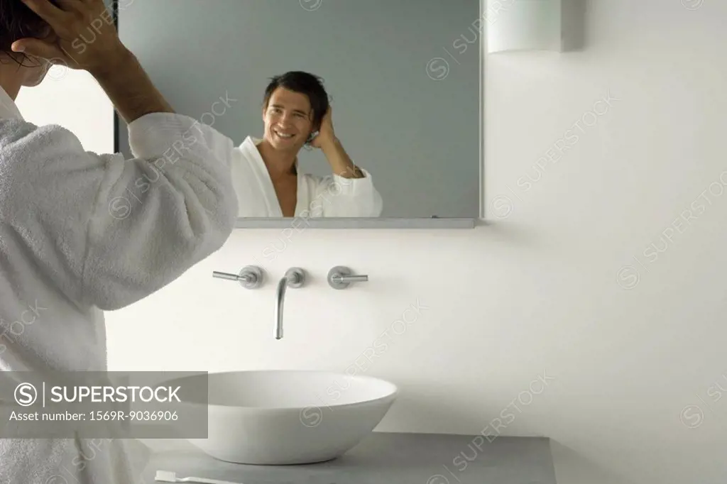 Man in bathrobe looking at self in bathroom mirror, hand in hair