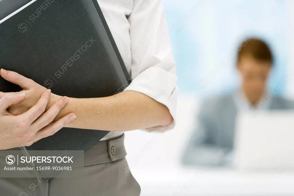 Professional woman holding binder, cropped view, male colleague in background