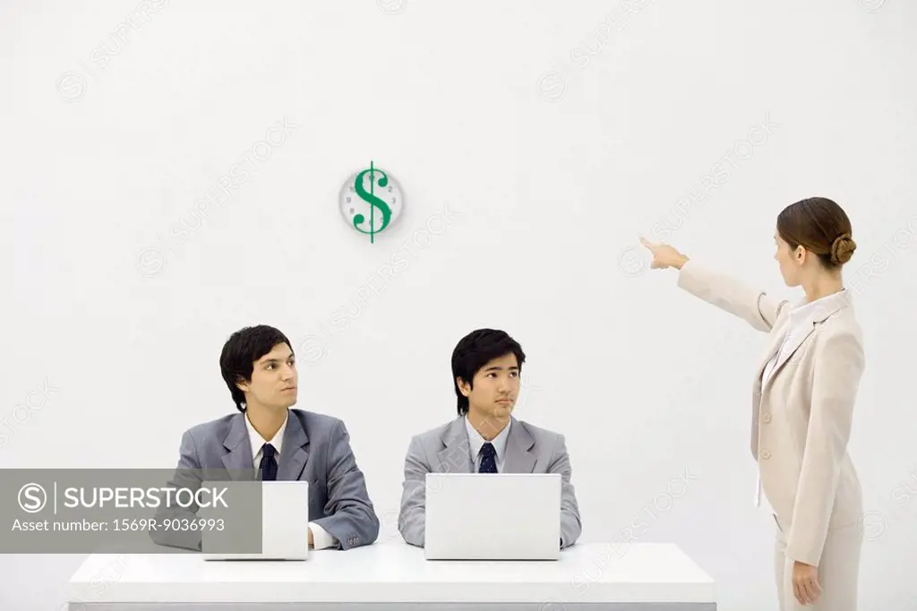 Two male office workers sitting at desk, female supervisor pointing at clock with dollar sign