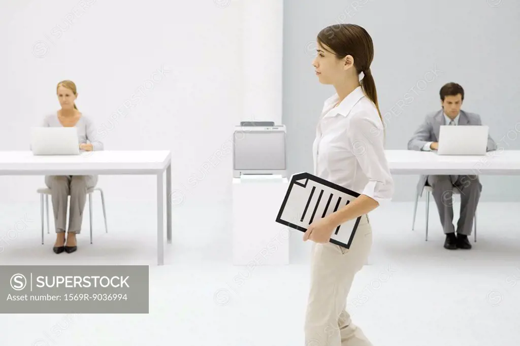 Female office worker carrying document under arm, colleagues working in background