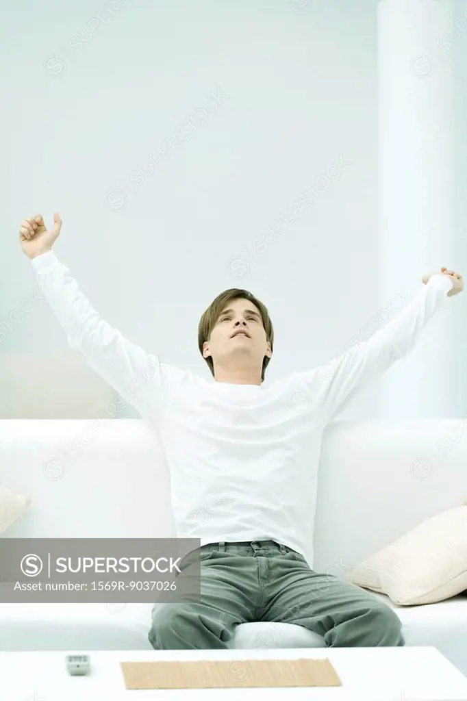 Young man sitting on couch with arms raised