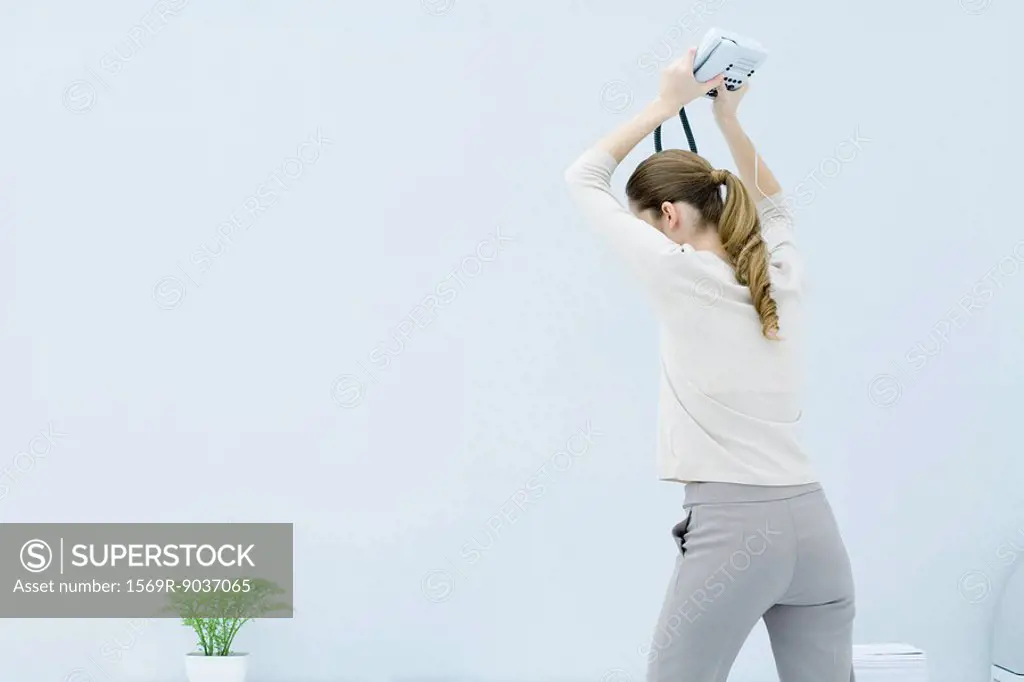 Young woman holding landline phone above her head ready to smash it on the ground, rear view