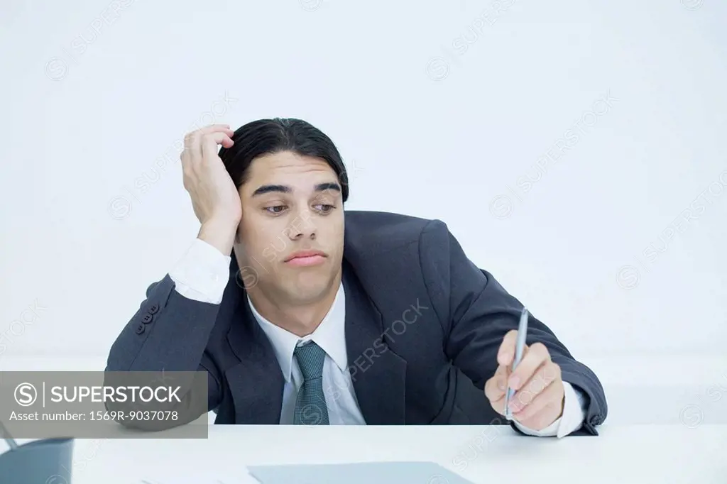 Young businessman slouching at desk, holding head and looking at pen with bored expression