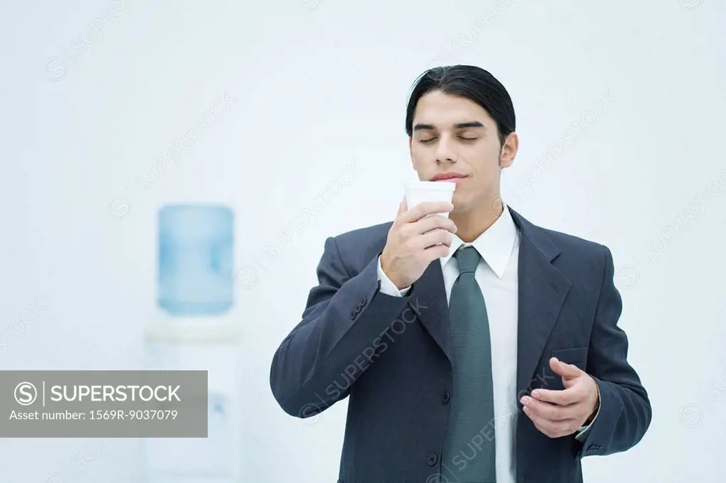 Businessman holding and smelling disposable cup with eyes closed, water cooler in background