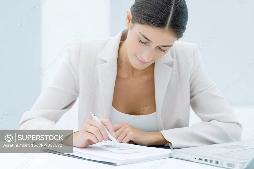 Young businesswoman using pen to enter numbers on calculator, looking down