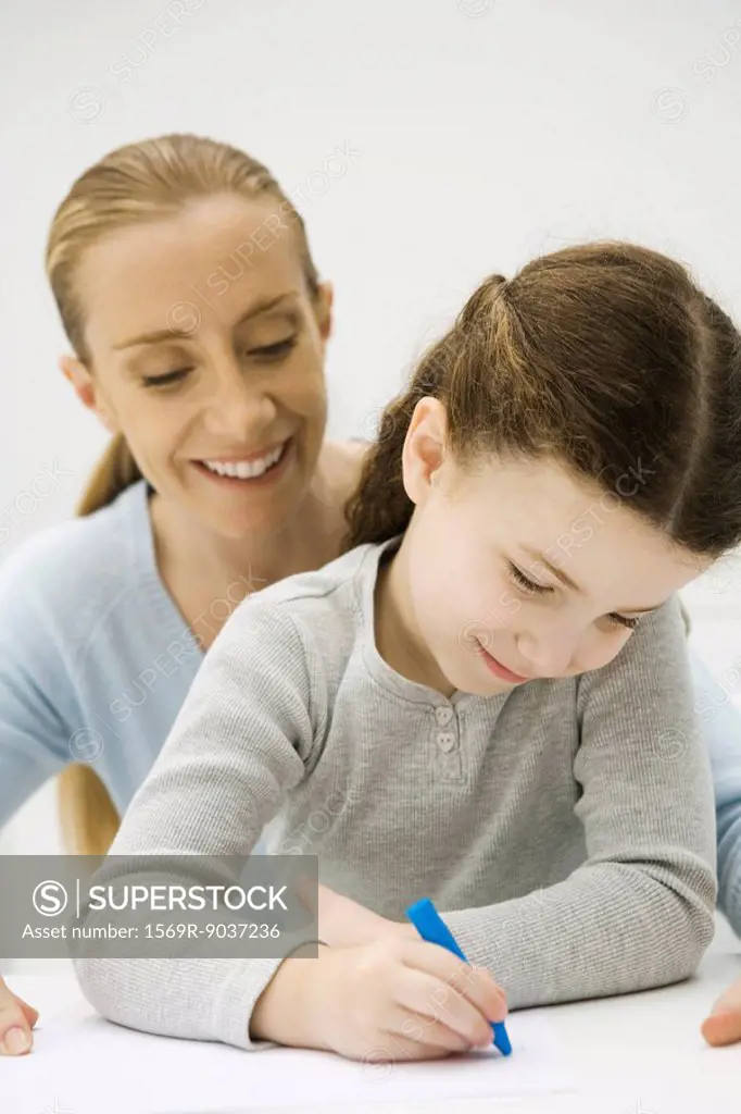 Girl drawing with crayon, mother looking over her shoulder and smiling