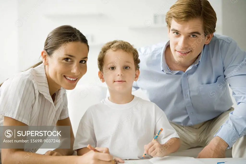 Little boy sitting with parents, drawing with pencil, all smiling at camera