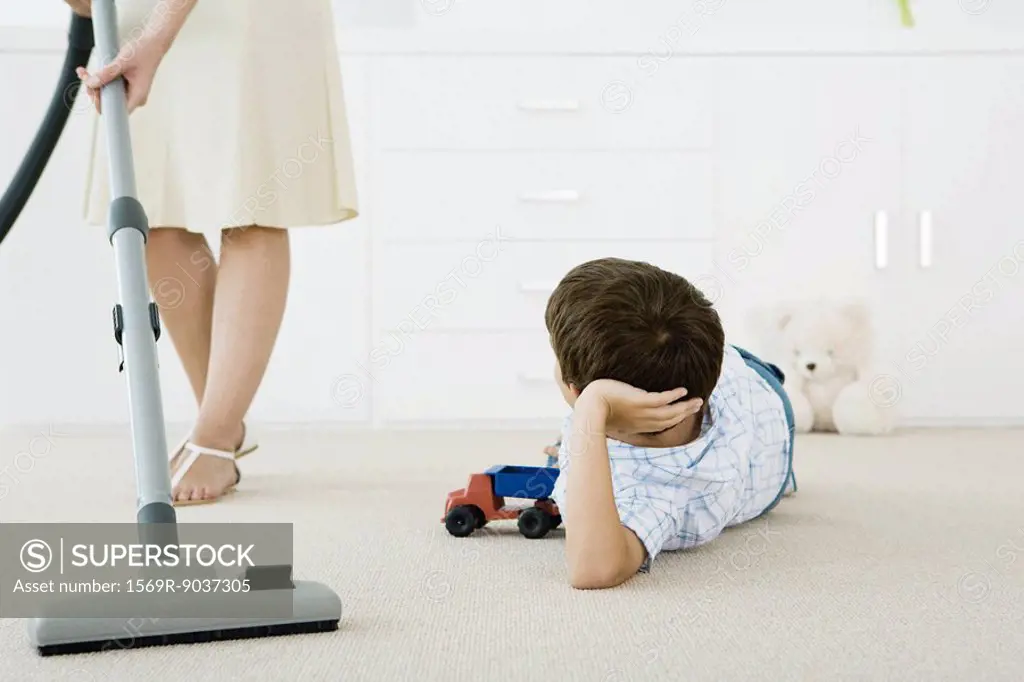 Boy lying on the ground with toys, looking up at his mother vacuuming around him