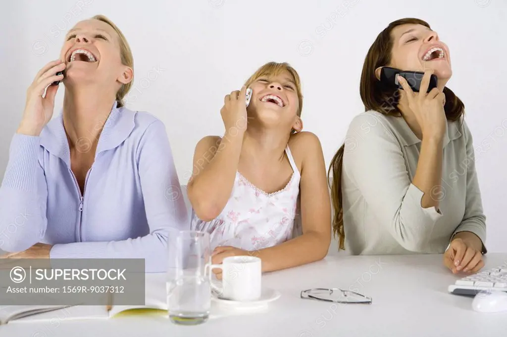 Two women and a preteen girl sitting at table, each using cell phone, all laughing and looking up