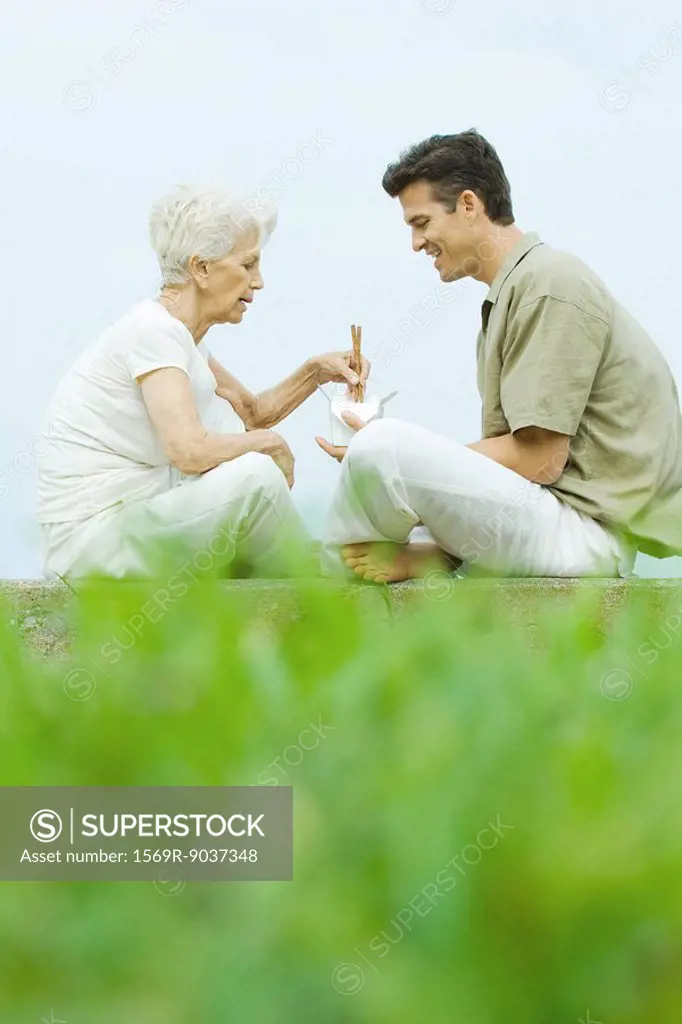 Senior woman with adult son, sitting in park sharing takeout food with chopsticks