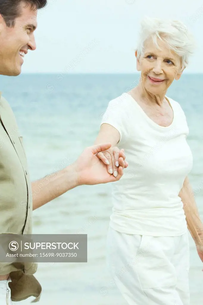 Senior woman walking hand in hand with adult son by water, smiling at each other