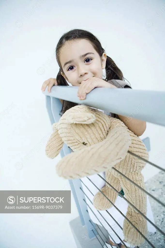 Little girl standing at rail holding teddy bear, smiling at camera